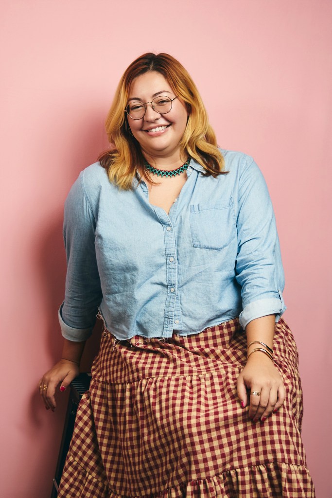 Light-skinned person with red and blonde hair leaning back on a stool, wearing a denim long-sleeved shirt and a long, flowy red gingham skirt.